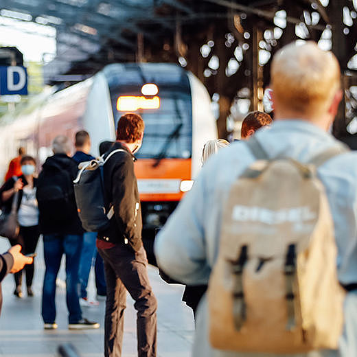 Mehrere Menschen warten an einem Bahngleis, an dem ein Zug einfährt.